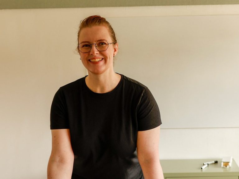 Katharina im Unterricht Katharina trägt eine Brille und ein schwarzes T-Shirt und steht vor einem White Board.